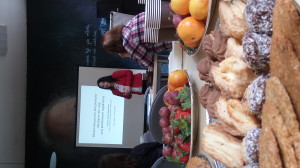 Zdjęcie przedstawia na pierwszym planie zbliżenie stołu z ciasteczkami i owocami, na drugim planie widać osobę siedzącą i słuchającą stojącej przy stojącym ekranie wykładowczyni