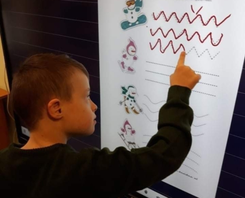 Zdjęcie chłopca piszącego szlaczki palcem po tablicy interaktywnej