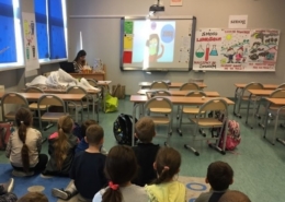 Zdjęcie przedstawia dzieci siedzące na podłodze w klasie szkolnej i oglądające prezentację na ekranie naściennym