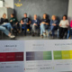 Zdjęcie przedstawia spotkanie grupy osób siedzących w kręgu na krzesłach w jasnym pomieszczeniu z ciemną ścianą w tle. Na pierwszym planie widoczna jest karta edukacyjna trzymana w ręku, zawierająca trzy kolorowe sekcje zatytułowane: „Zmaganie”, „Unikanie” i „Współpracowanie”. Każda sekcja opisuje inne podejścia do trudności w relacjach i współpracy – od konfliktu i krytyki, przez wycofanie, po budowanie porozumienia. W tle, nieco rozmyci, widoczni są uczestnicy spotkania, którzy biorą udział w warsztatach lub szkoleniu.
